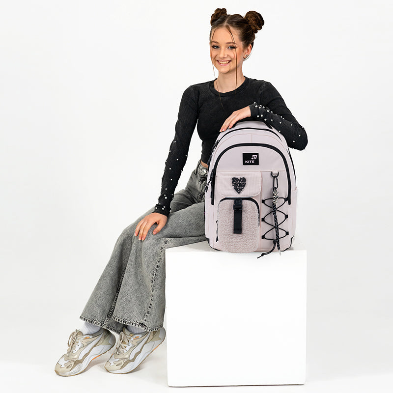 Modelka nastolatka siedząca obok jasnoróżowego plecaka Kite, co pozwala ocenić jego wielkość i styl.